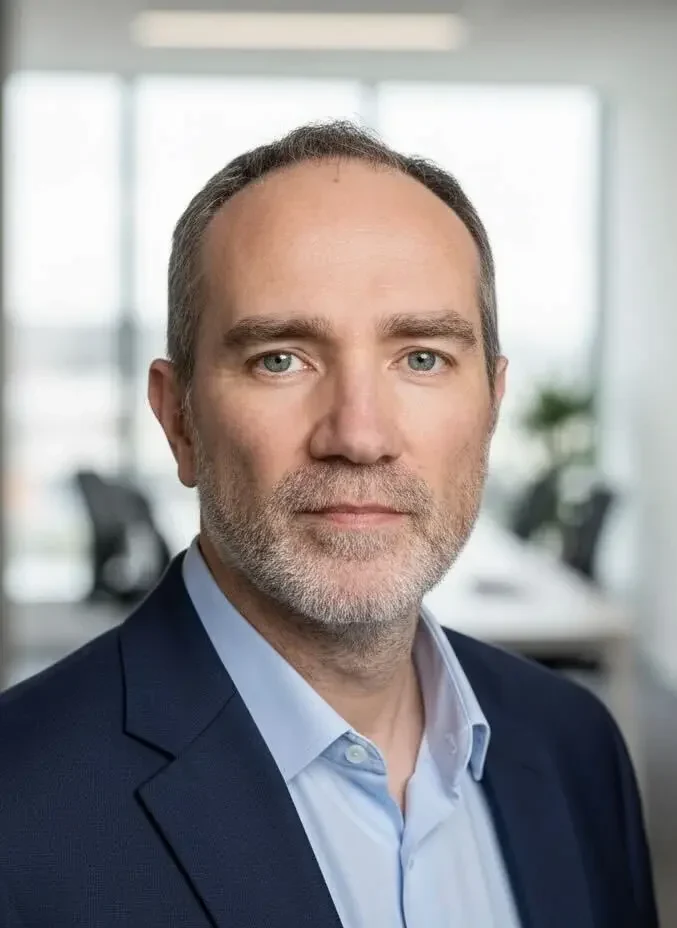 A middle aged man with short gray hair and a beard, wearing a navy suit and light blue shirt, stands in a modern office setting.