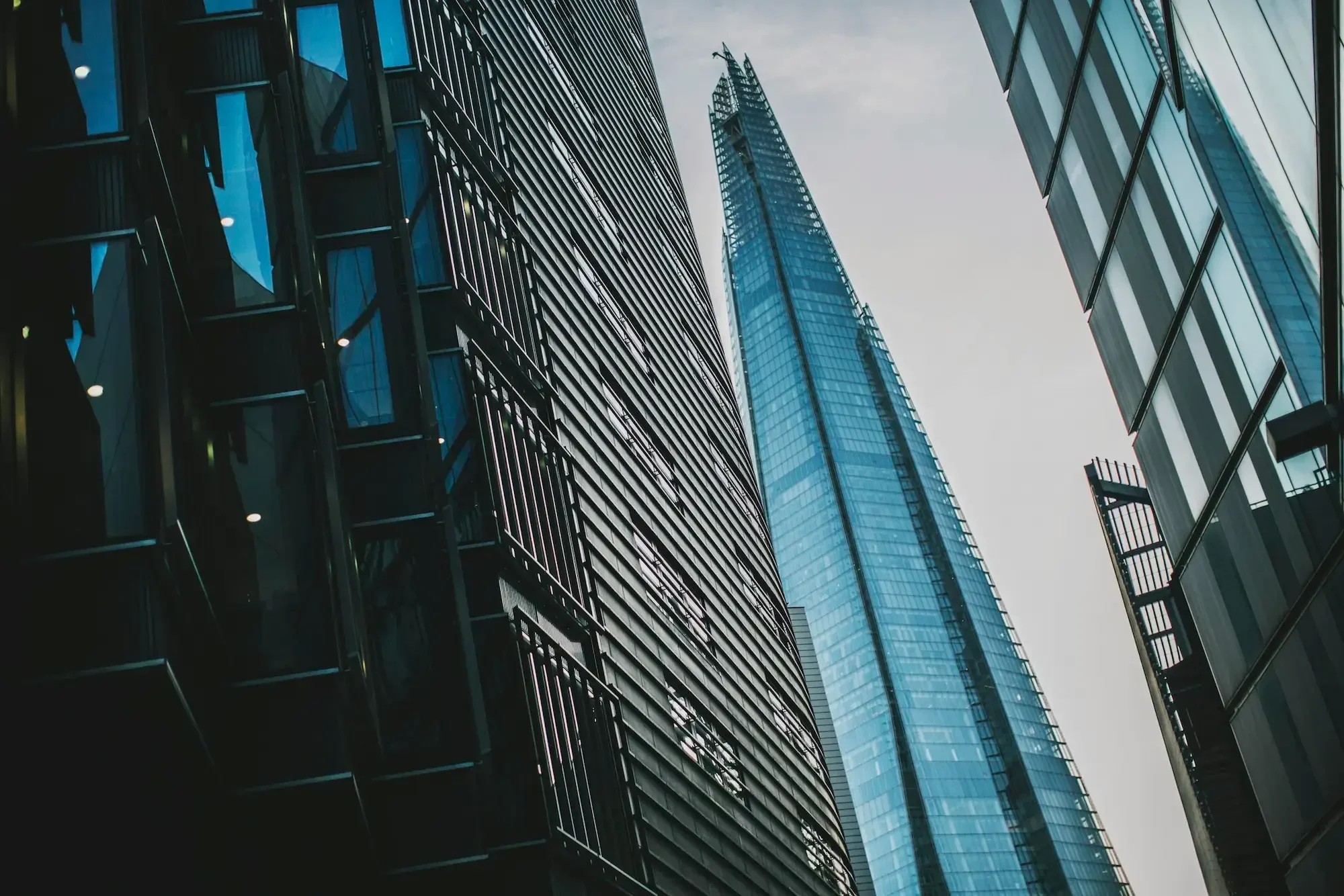 Tall modern skyscrapers with glass facades, including the pointed shard building, viewed from street level against a cloudy sky.