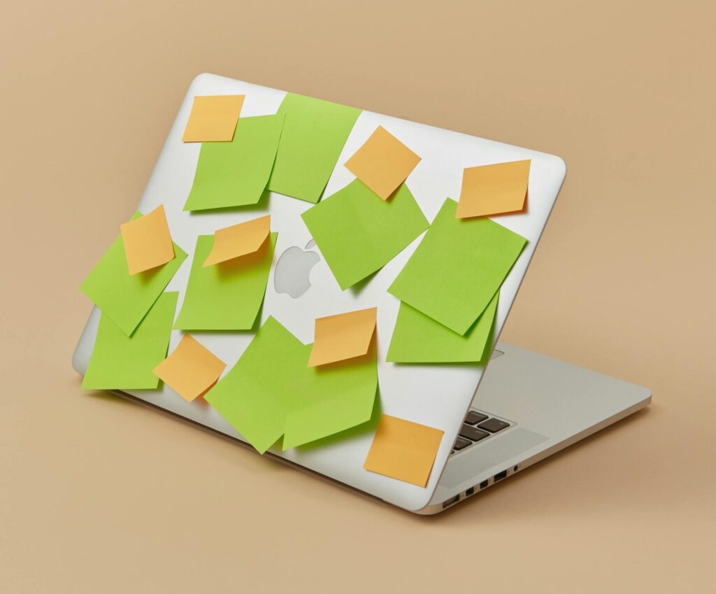 A laptop with its top panel covered in vibrant green and yellow sticky notes, isolated on a beige background.