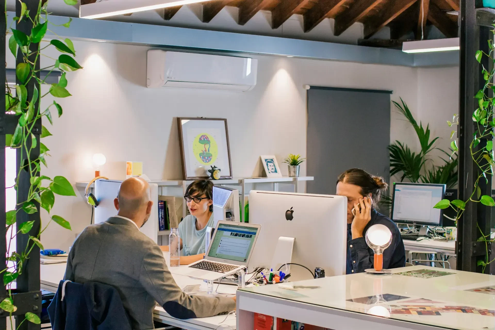 Three people work at desks with computers in a modern office space, surrounded by plants and office supplies. one person is on the phone, while others are engaged in conversation.
