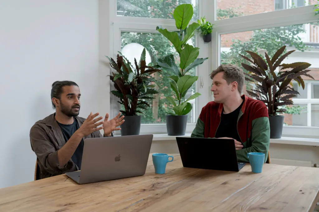 Two people sit at a wooden table with laptops and coffee mugs, having a discussion. There are large green plants and big windows in the background, letting in natural light.