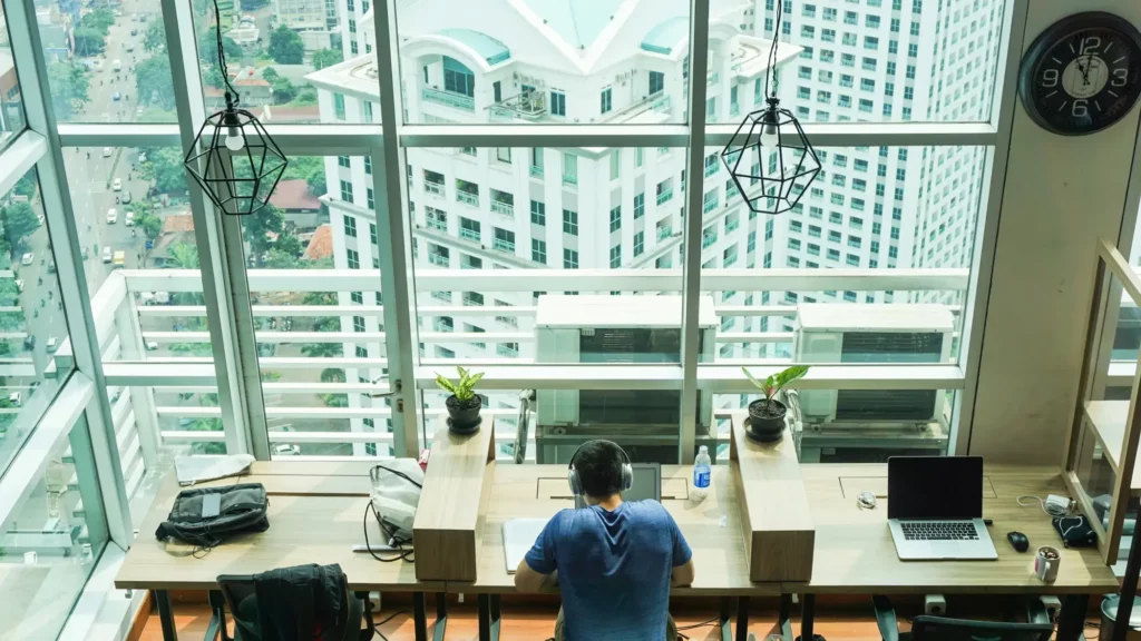 Person working at a wooden desk in a modern office with a city view. Three laptops and potted plants are on the desk. Large windows overlook urban buildings.