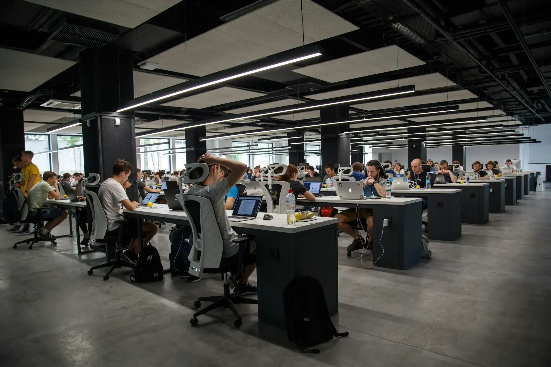 Large open plan office with many people working at desks on computers, under bright ceiling lights, in a modern setting with gray floors and black columns.