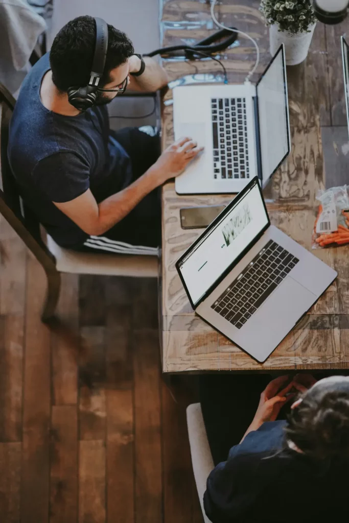 Two people work at a wooden table with laptops, headphones on. A smartphone and snacks are nearby. View from above.