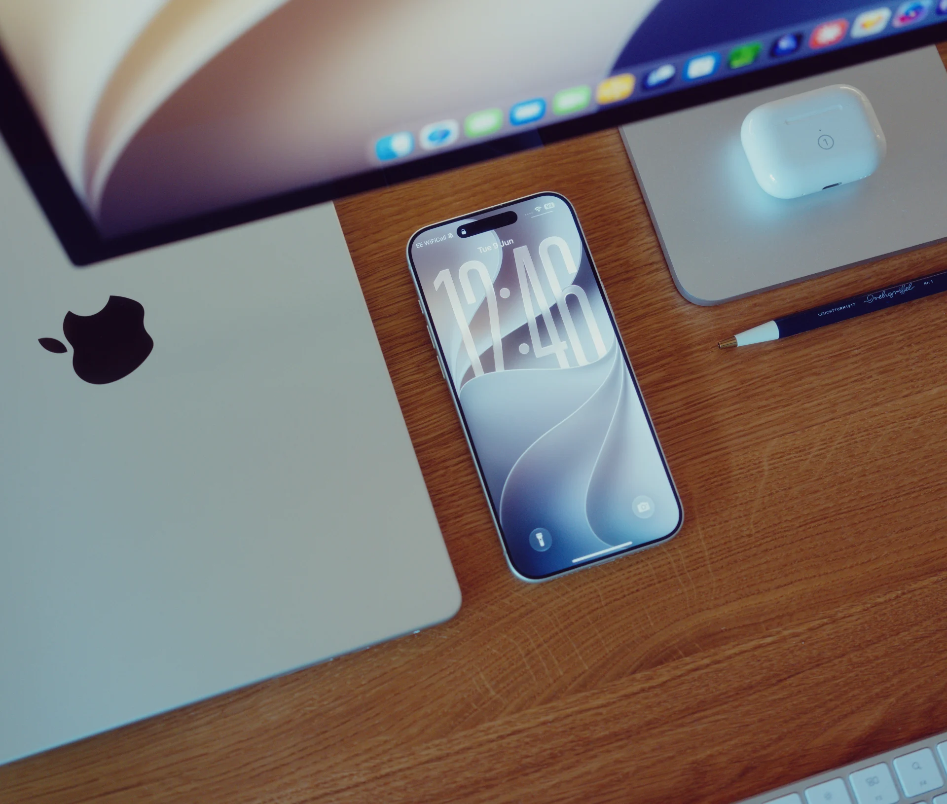 A smartphone displaying the time rests on a wooden desk next to a closed laptop, a computer monitor, and a wireless earbud case.