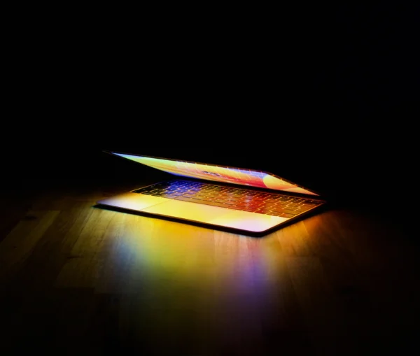 A partially open laptop sits on a wooden surface, casting a vibrant glow onto the table against a dark background as it prepares to restart Mac.