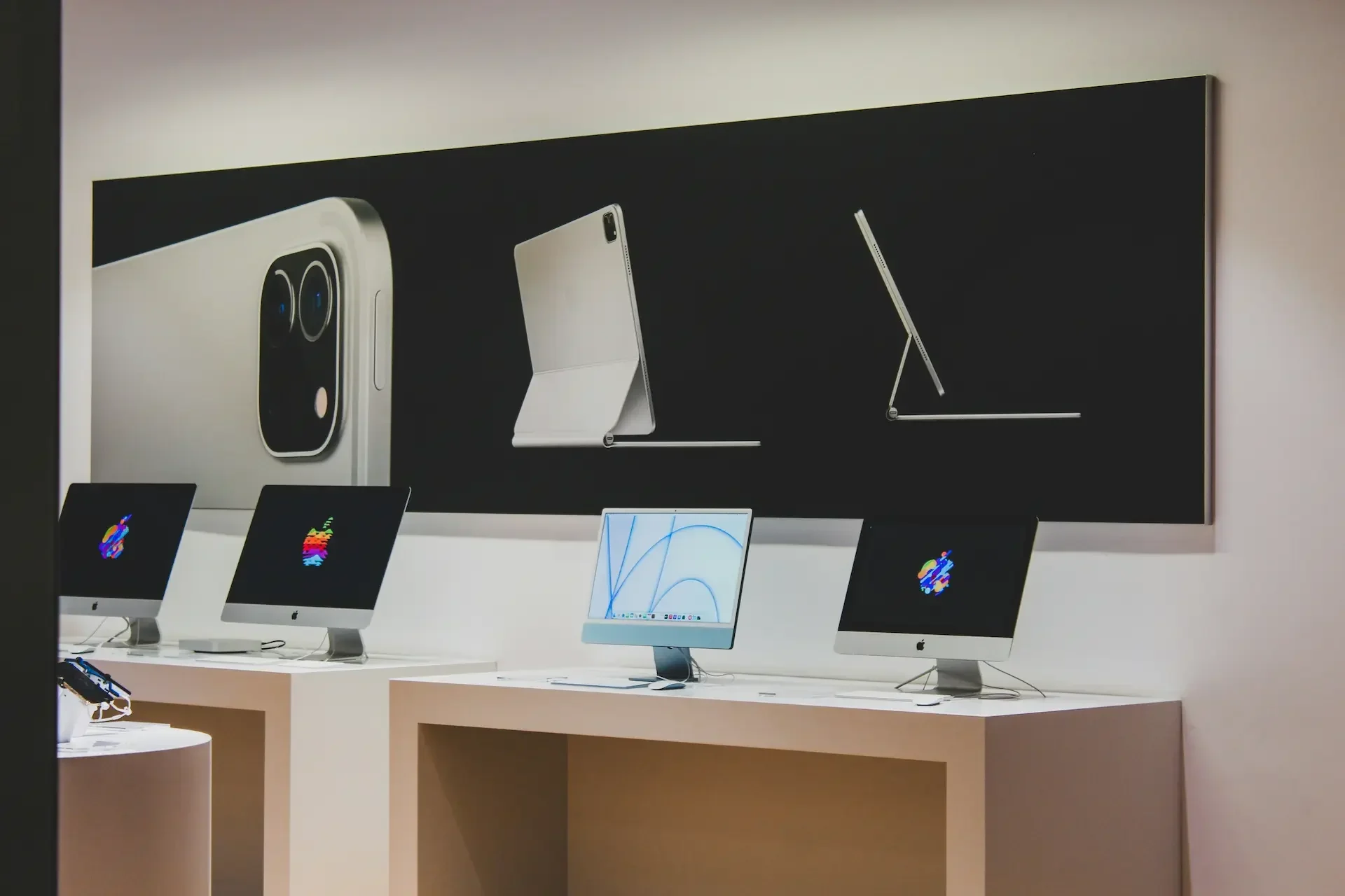 Four apple desktop computers are displayed on tables beneath a wall poster featuring images of an ipad, ipad keyboard, and close up of an ipad camera—showcasing the benefits of mac it support without hidden cost concerns.