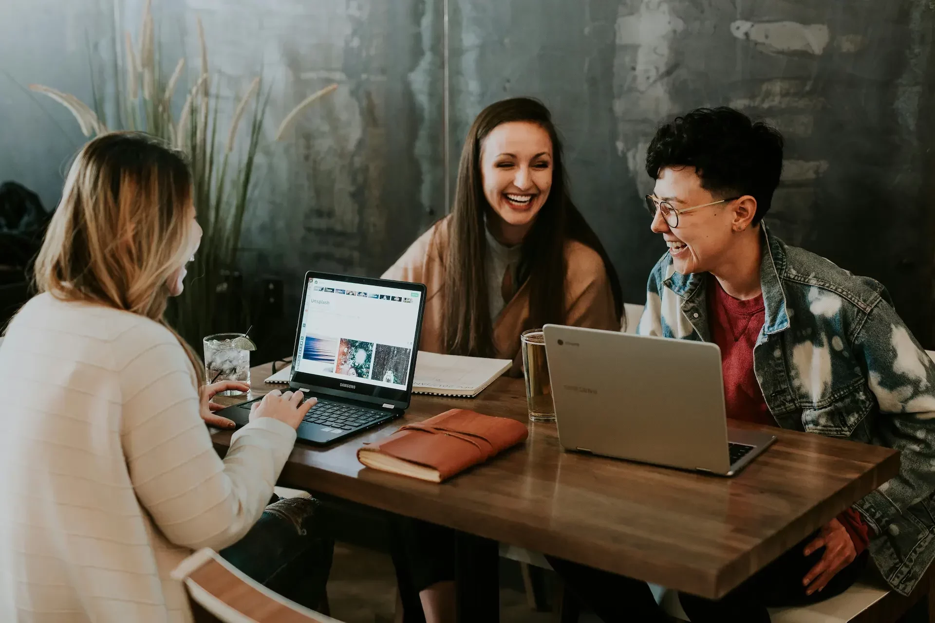 Three people sit at a wooden table with laptops, notebooks, and a glass of water, smiling and engaging in conversation about cyber essentials in a casual indoor setting.