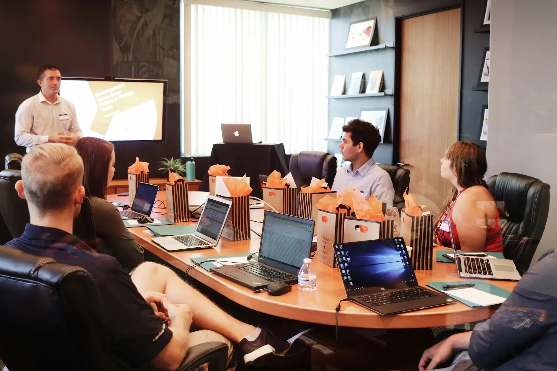 A group of people sit around a conference table with laptops, intently watching a man give a presentation on governance and risk management displayed on the large screen.