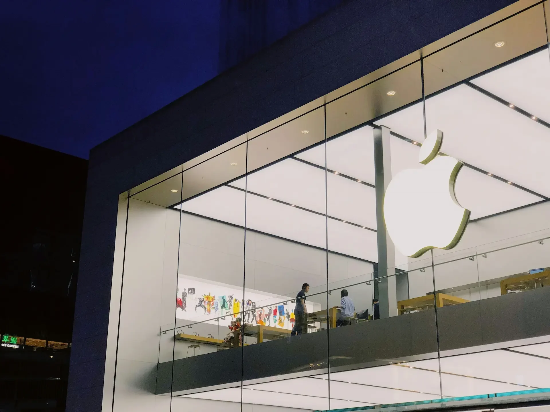 A two story apple store with large glass windows, a brightly lit interior, and a prominent white apple logo on the upper facade stands as an icon of innovation, adapting to eu regulations like the digital markets act.