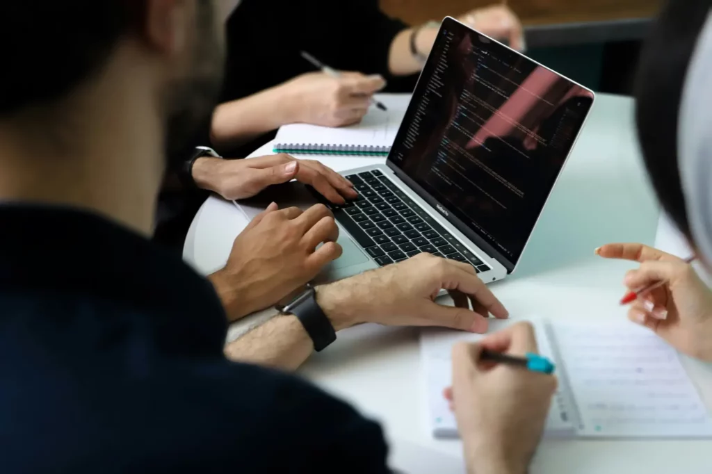 Three people are gathered around a laptop showing code, taking notes and discussing cyber security strategies.