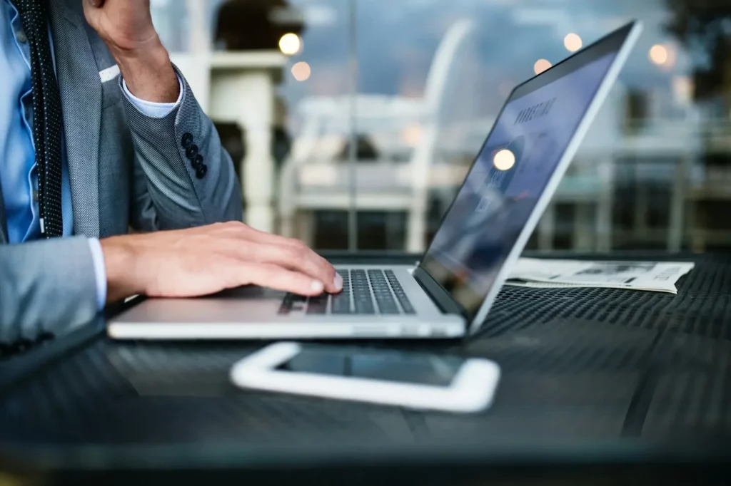Person in a suit using a laptop on a table with a smartphone and a newspaper nearby.