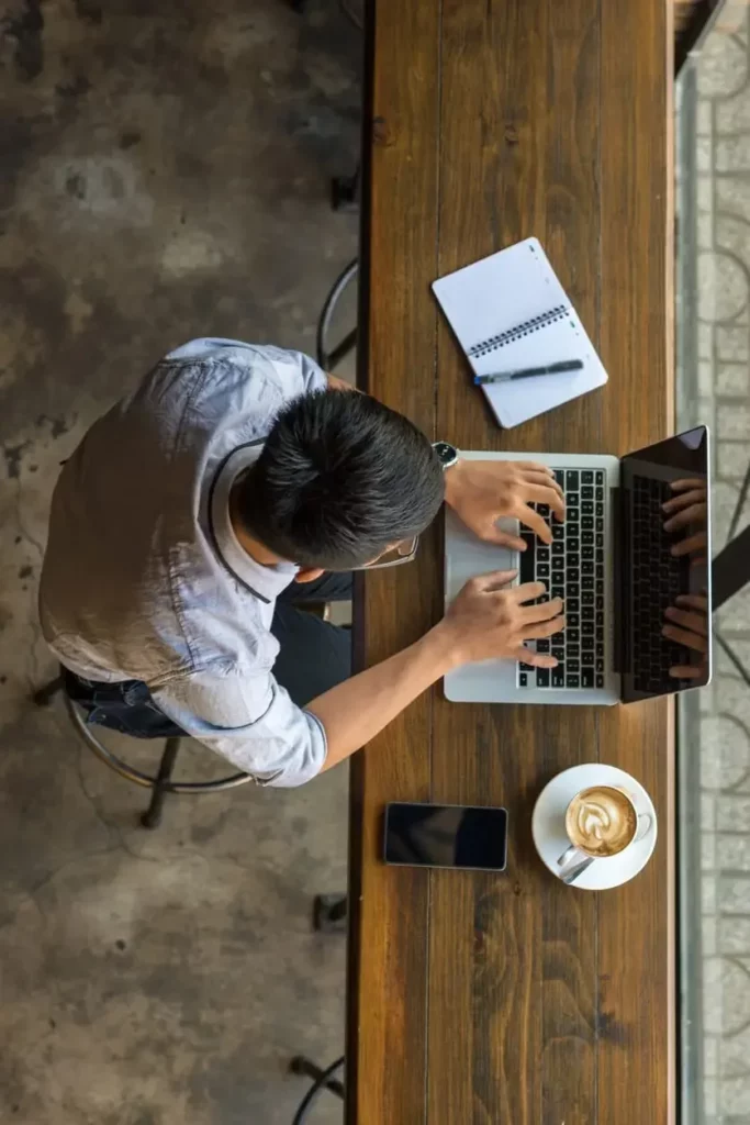 A person focused on cyber security types diligently on a laptop at a wooden table, accompanied by a notebook, smartphone, and a nearby cup of latte. The scene is captured from above.