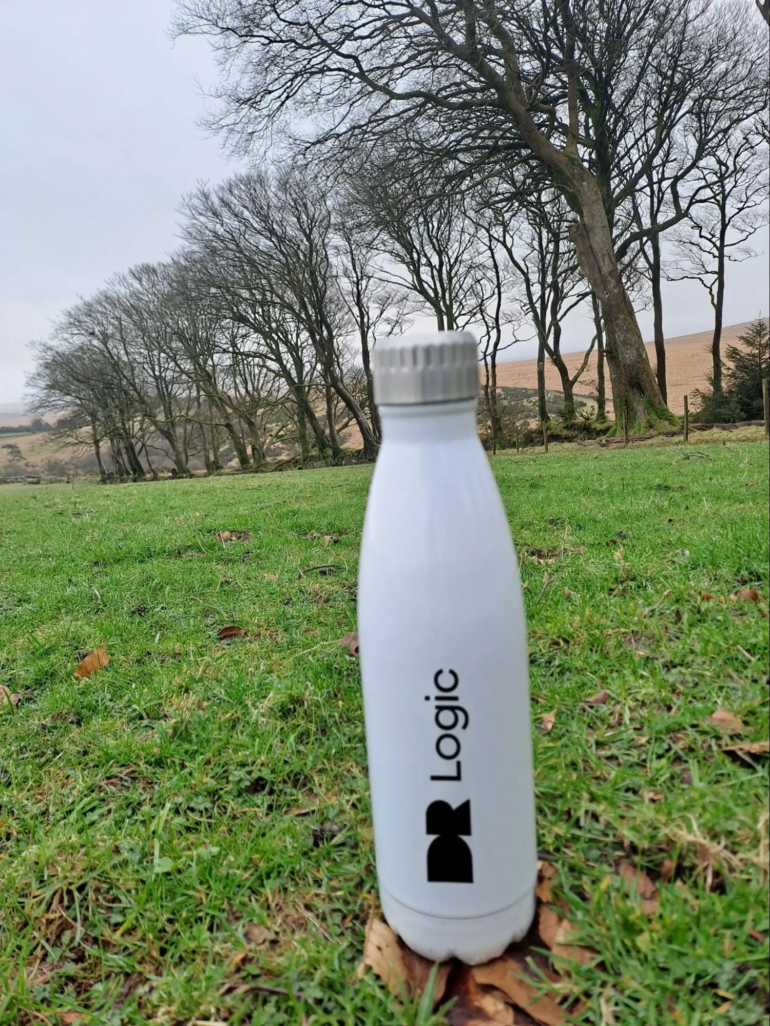 A white reusable water bottle labelled "DR Logic" stands on green grass with scattered brown leaves. In the background, leafless trees line the Dartmoor horizon under a cloudy sky, evoking a sense of sustainability and nature.