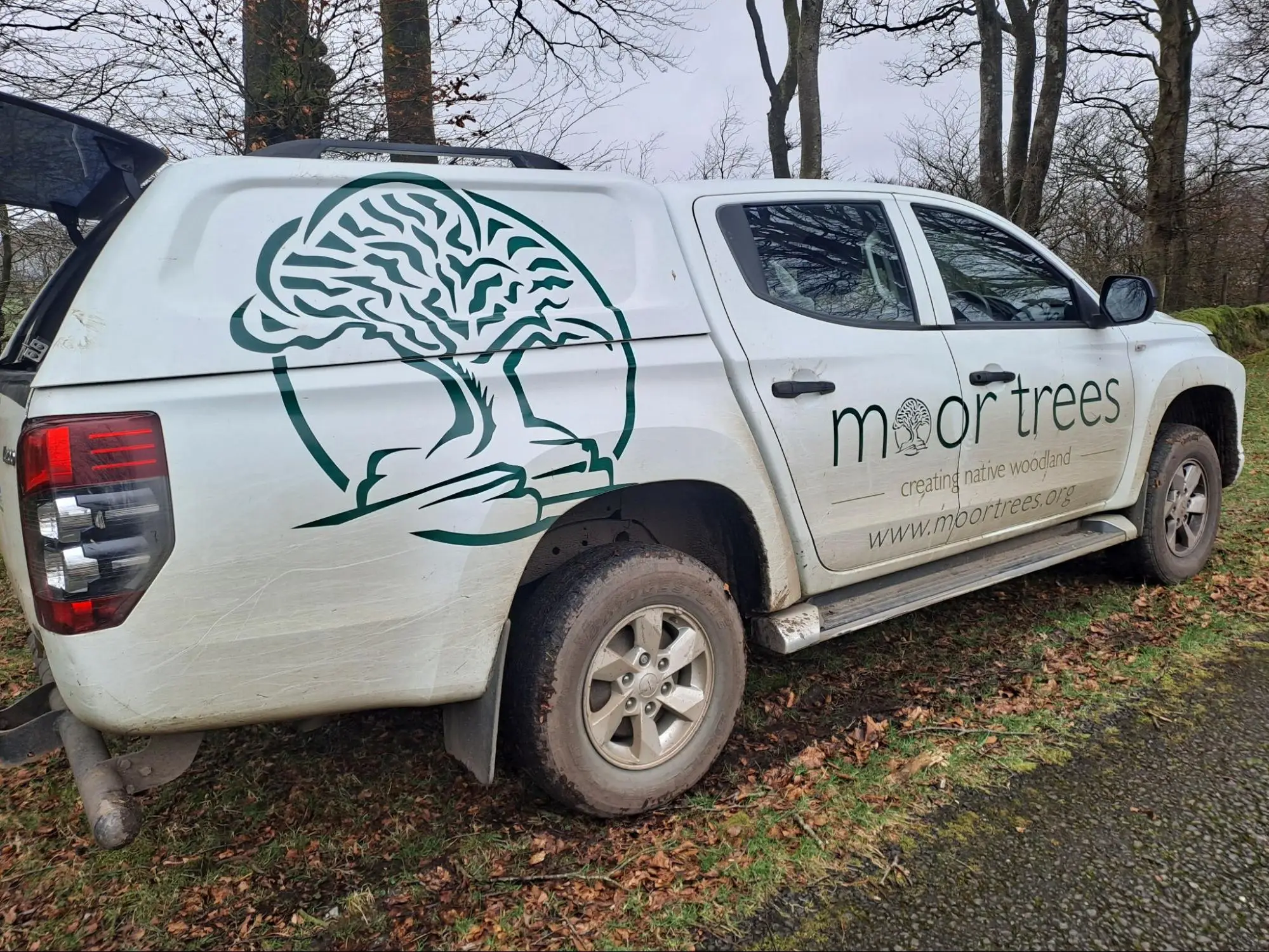 A white pickup lorry parked on grass, featuring a large tree graphic and the text "Moor Trees – creating native woodland in Dartmoor – www.moortrees.org" on its side.
