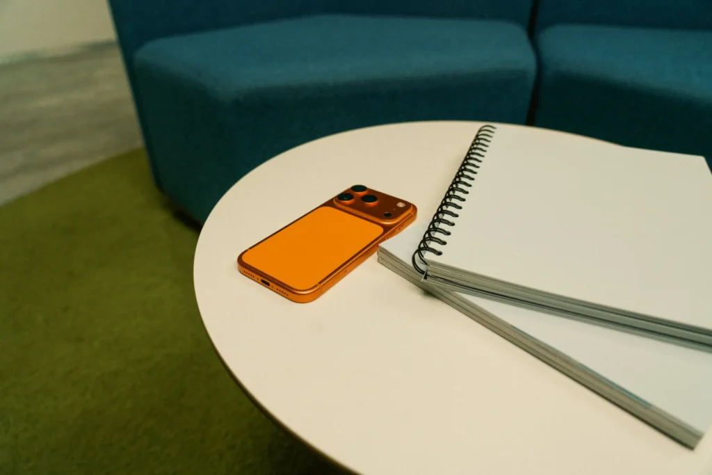 An orange mobile phone and a spiral notebook are placed on a round white table next to a blue sofa and green carpet, creating an organised space ideal for efficient iPhone management.