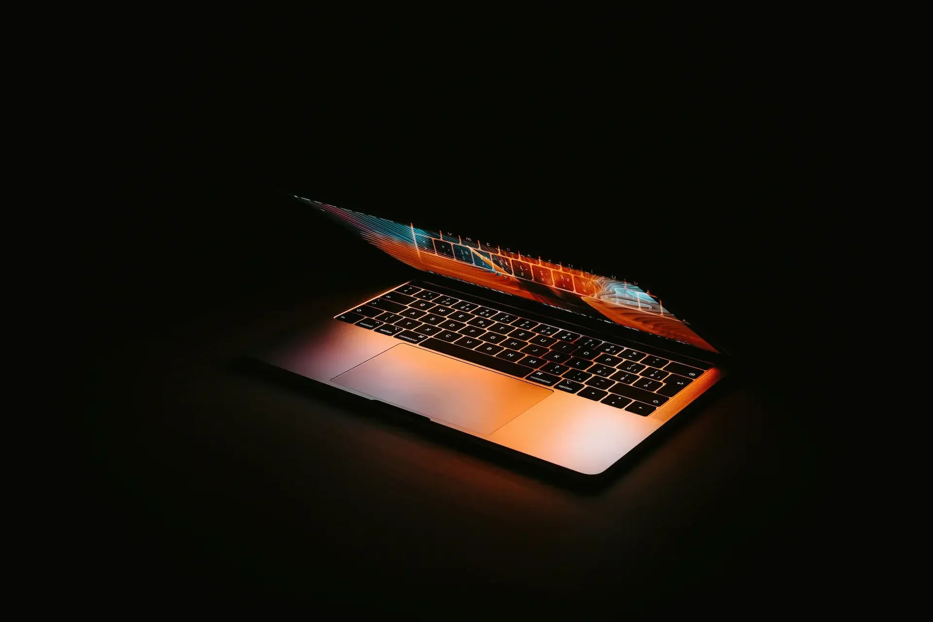 A partially open laptop with an illuminated screen running macos ventura sits in a dark environment, casting a warm glow on the keyboard and surrounding area.