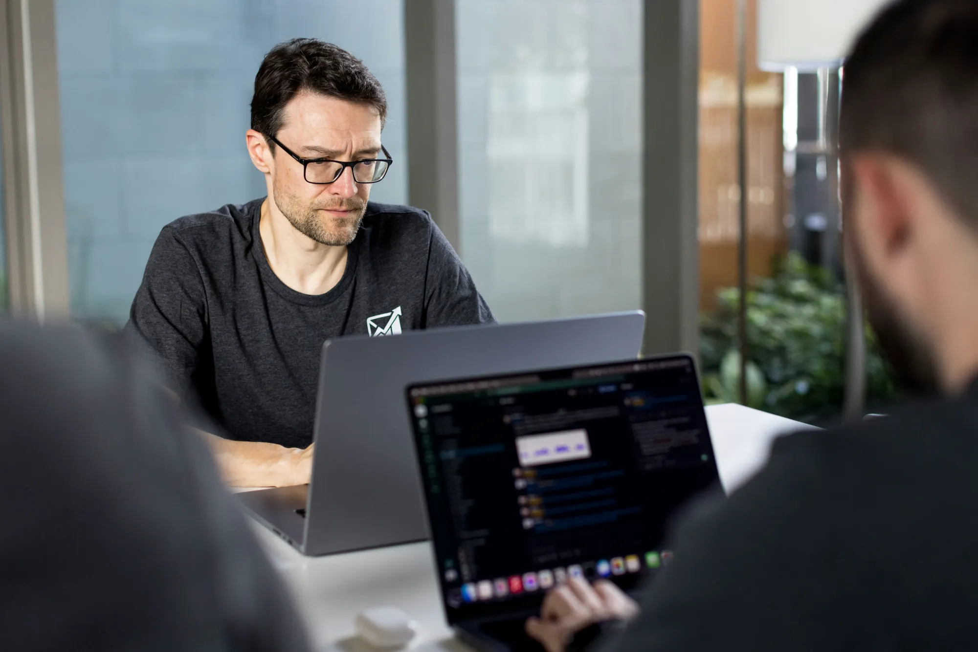 A man in glasses works on a laptop at a desk, focused on cyber security tasks, whilst another person with a laptop is in the foreground. They appear to be in a modern, bright office space with large windows and indoor plants.