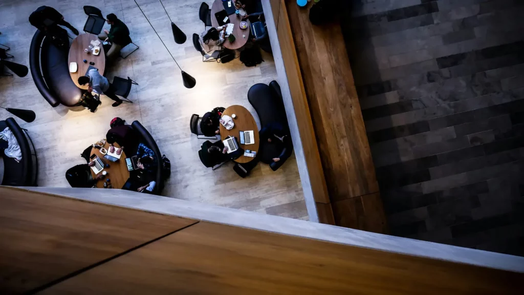 High-angle view of the team sitting at oval tables with laptops and papers in a modern, indoor setting. The space features wooden and tile flooring and hanging lights.