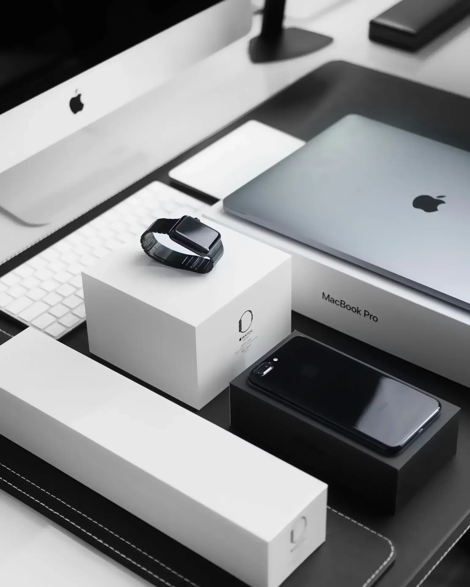 A macbook pro, apple watch, and iphone in their boxes are arranged on a desk with a keyboard and an imac in the background—perfect for any apple consultants network professional.