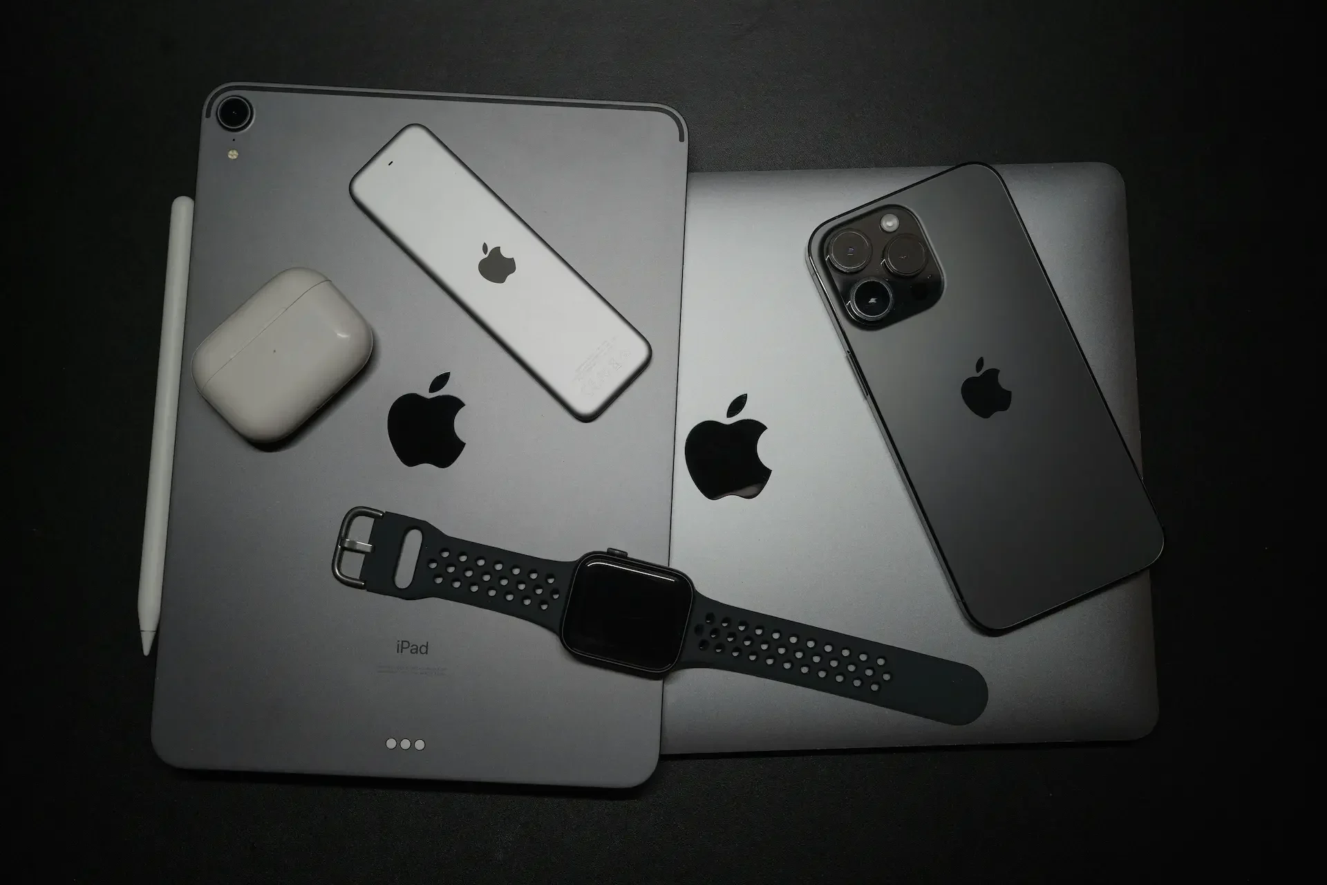 A collection of apple devices, perfect for streamlined apple device management, including an ipad, two macbooks, an iphone, airpods, an apple tv remote, an apple pencil, and an apple watch on a black surface.