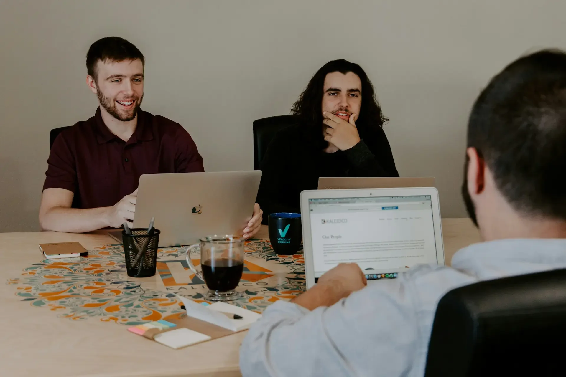 Three people sit at a table with laptops, notepads, and drinks, engaged in a work meeting or discussion that appears to focus on quarterly business reviews.