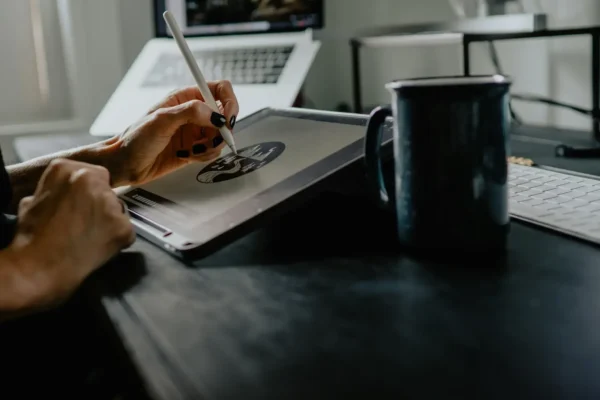 A person from a uk design studio uses a stylus to draw on a tablet at their desk, with a mug, keyboard, and open laptop—perhaps troubleshooting designs with remote it support in the background.