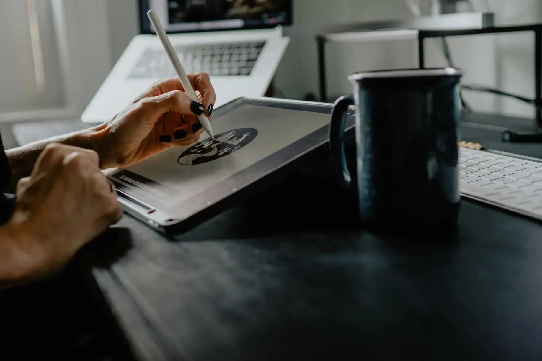 A person from a uk design studio uses a stylus to draw on a tablet at their desk, with a mug, keyboard, and open laptop—perhaps troubleshooting designs with remote it support in the background.