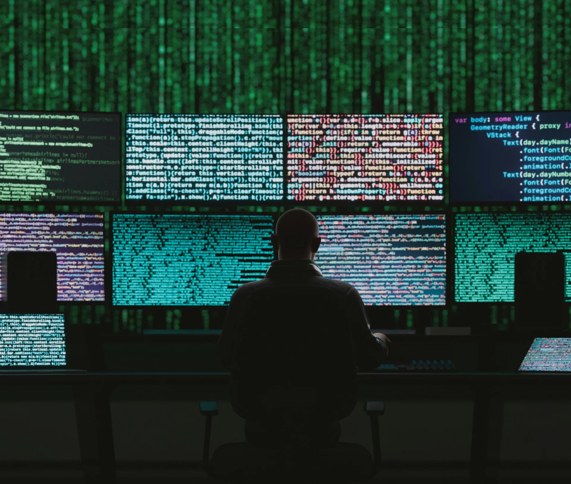 A person sitting in a dark room facing multiple computer monitors displaying cascading green lines of code.