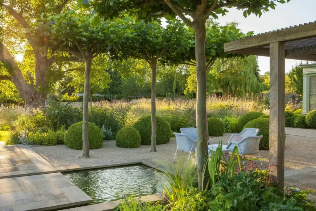 A serene garden scene designed by Marcus Barnett Studio features a small pond, four wicker chairs around a table, and manicured bushes under tall, leafy trees. A pergola and gravel pathway complete this harmonious retreat.