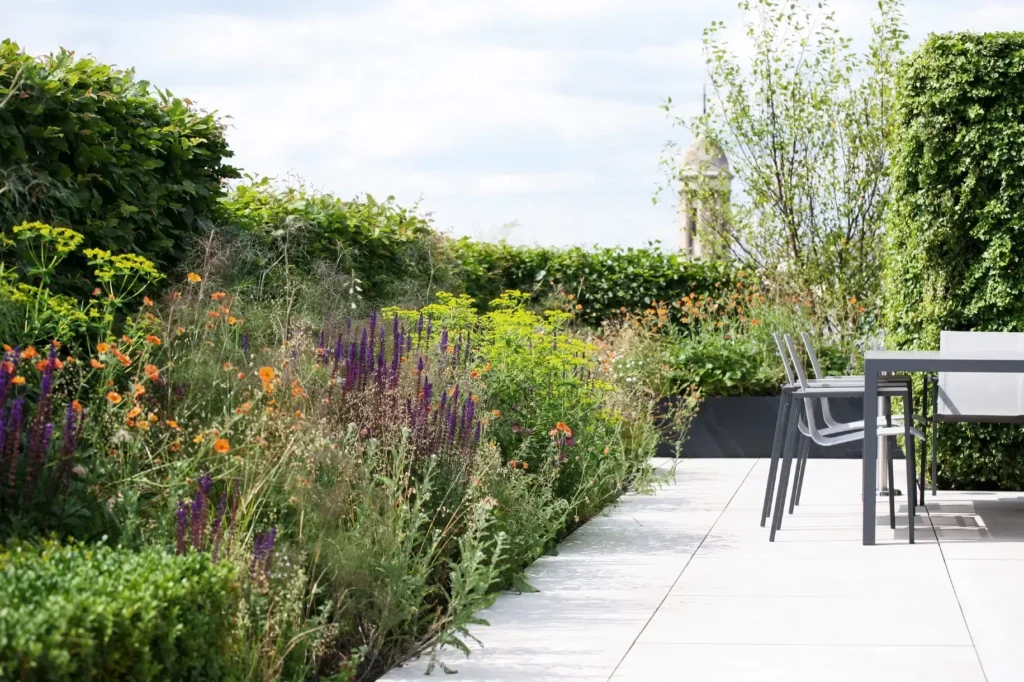 A modern patio with a dining table and chairs is expertly crafted by Marcus Barnett Studio, bordered by a lush garden teeming with colorful flowering plants and green shrubs, all set under a cloudy sky.