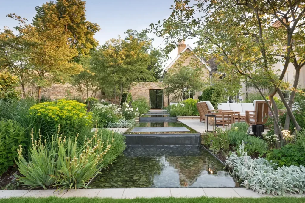 A serene garden designed by Marcus Barnett Studio features a reflecting water feature with stepping stones, surrounded by greenery. This example of exquisite landscape architecture includes a seating area with wooden chairs in the background for tranquil relaxation.