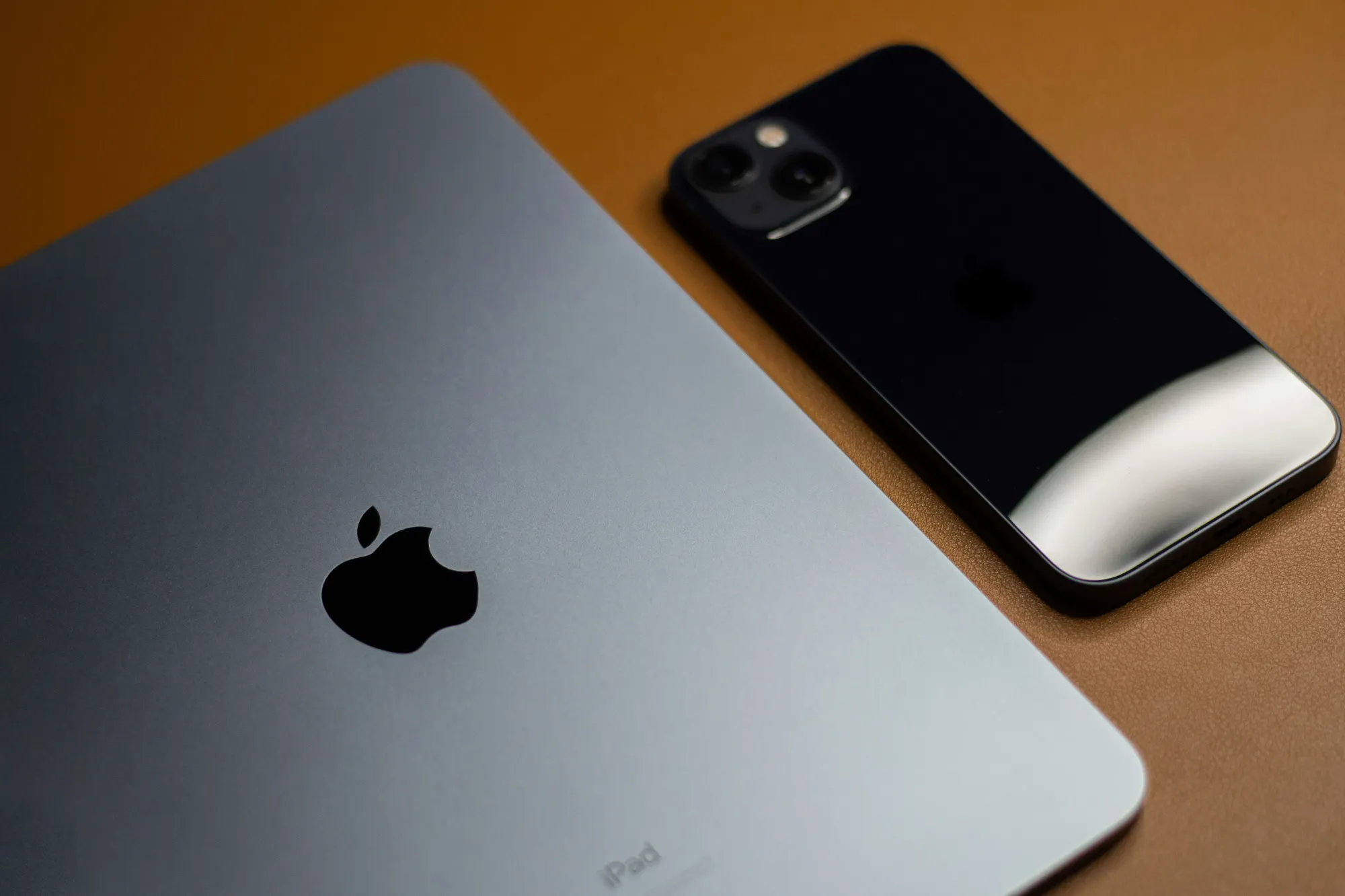 A close up of an Apple iPad and iPhone placed on a brown surface, both showing the Apple logo, highlighting seamless iPhone management and iPad management capabilities.