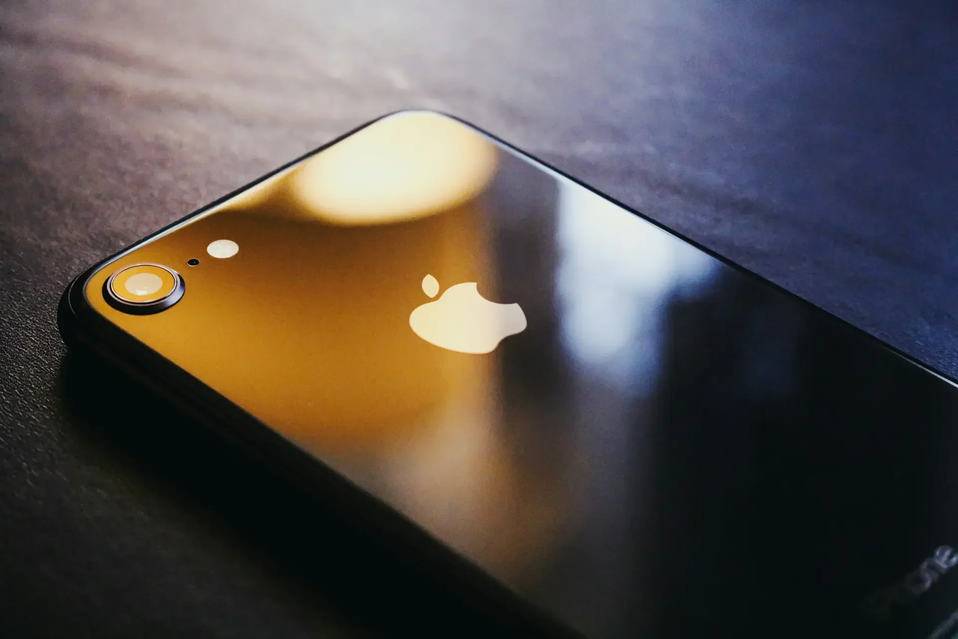 Close up of the back of a black iphone featuring the apple logo and camera lens, reflecting warm light on a dark surface—an essential device for business security and staying up to date with every urgent ios update.