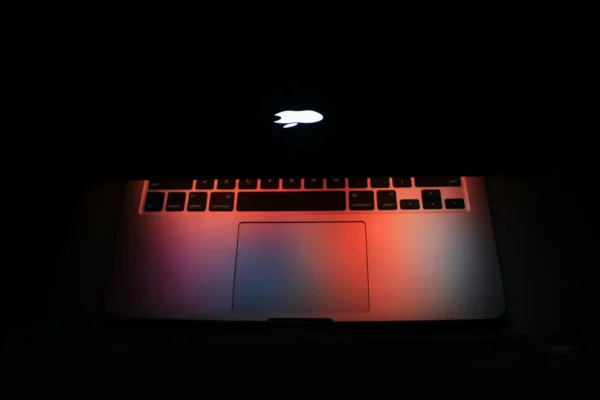 A partially closed macbook laptop with an illuminated apple logo casts red and blue light on the keyboard and trackpad in a dark environment, hinting at concerns around cyber breaches and the growing importance of a cybersecurity budget.