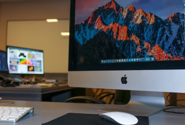 Close up of an apple imac with a mountain wallpaper on the screen, a white mouse and keyboard on the desk, and another monitor in the background—ideal for business it support environments.
