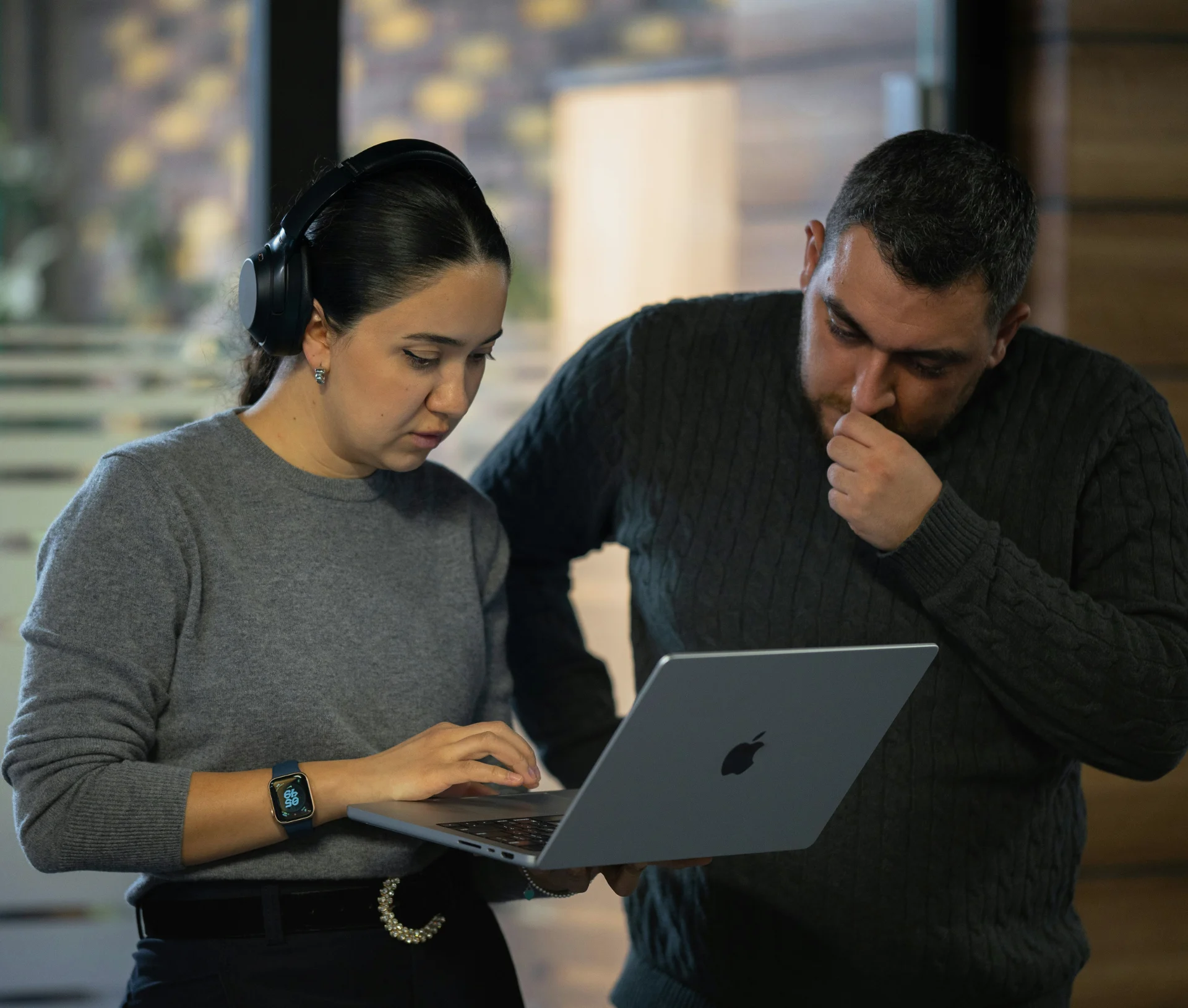 A woman wearing headphones and a man standing beside her looking at a laptop screen during an Apple managed service provider session.