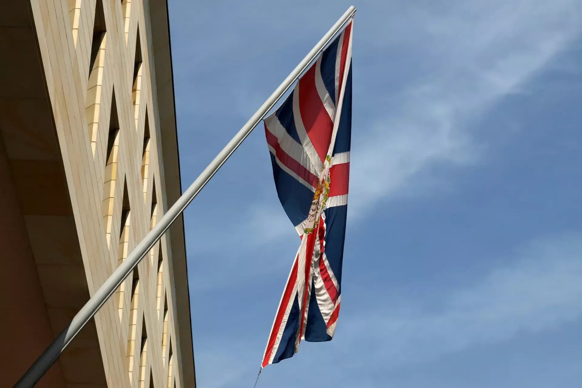 A torn and tattered british flag is flying on a flagpole beside a building against a partly cloudy blue sky, recalling recent news of the 23andme data breach.