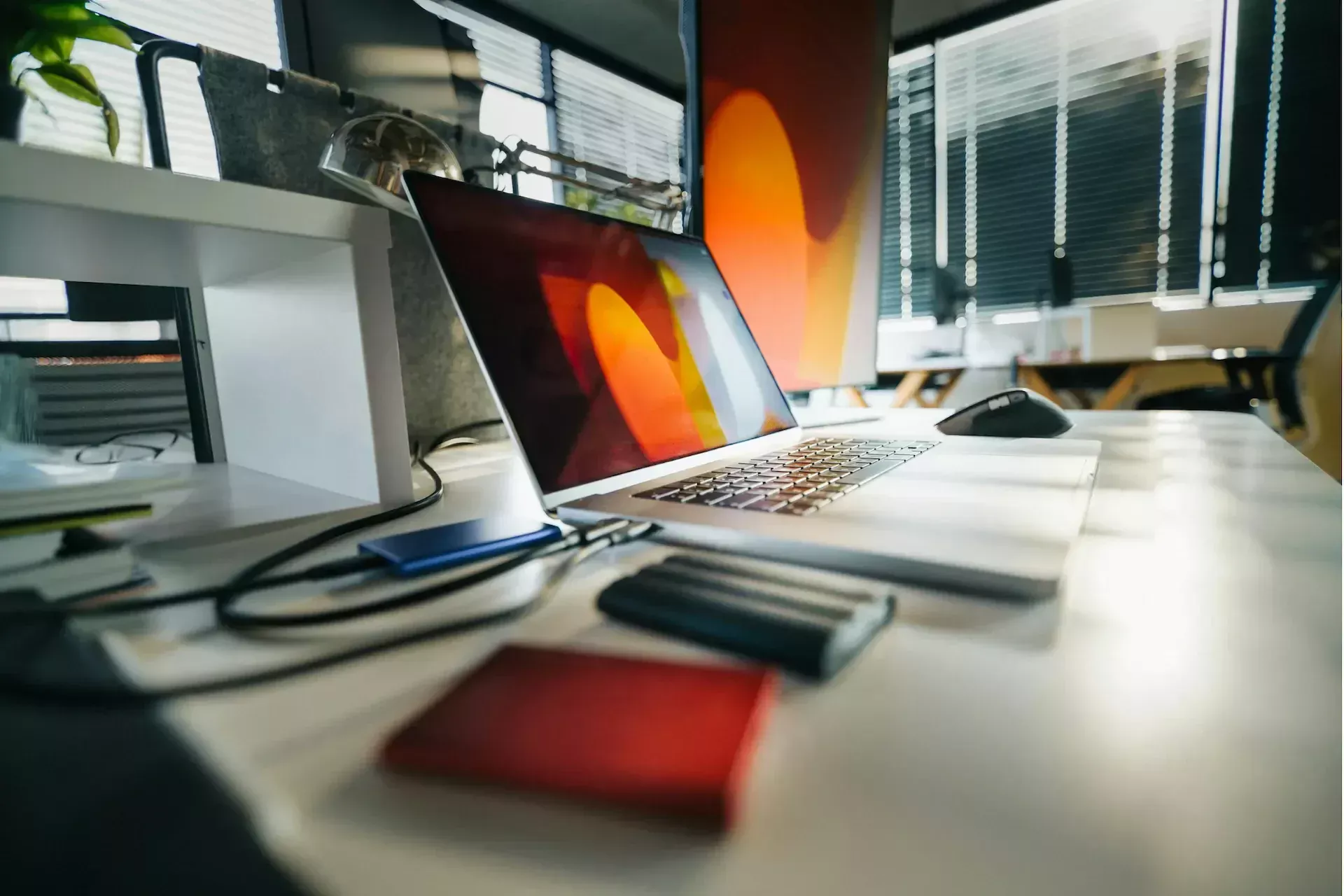 A laptop with a colorful screen sits on a desk alongside external hard drives, backup devices, and other office items in a modern workspace with window blinds—highlighting the importance of a business safety net.