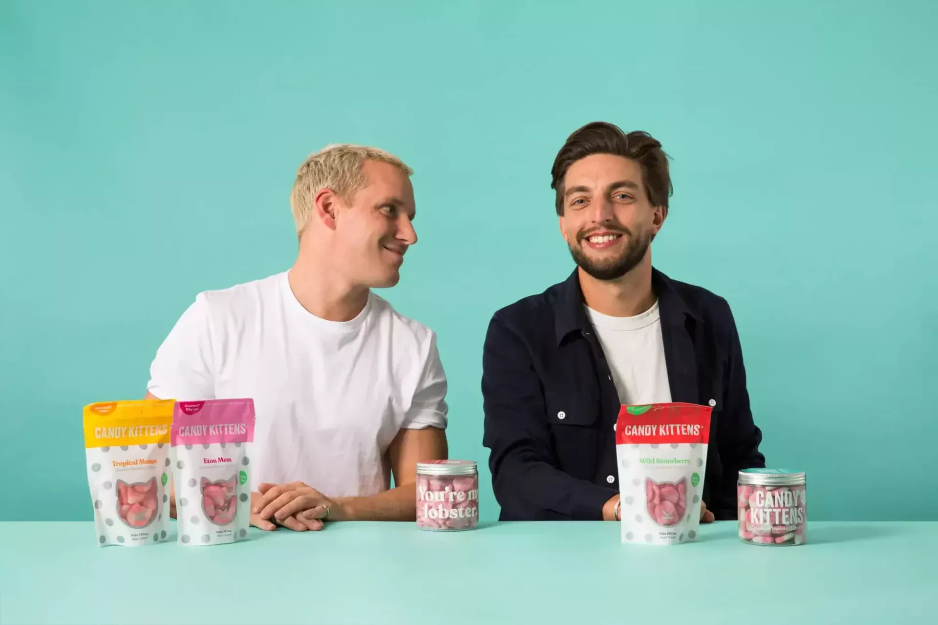 Two men are seated at a table with an array of Candy Kittens products displayed in front of them against a teal background, creating a scene as sweet as the candy itself.
