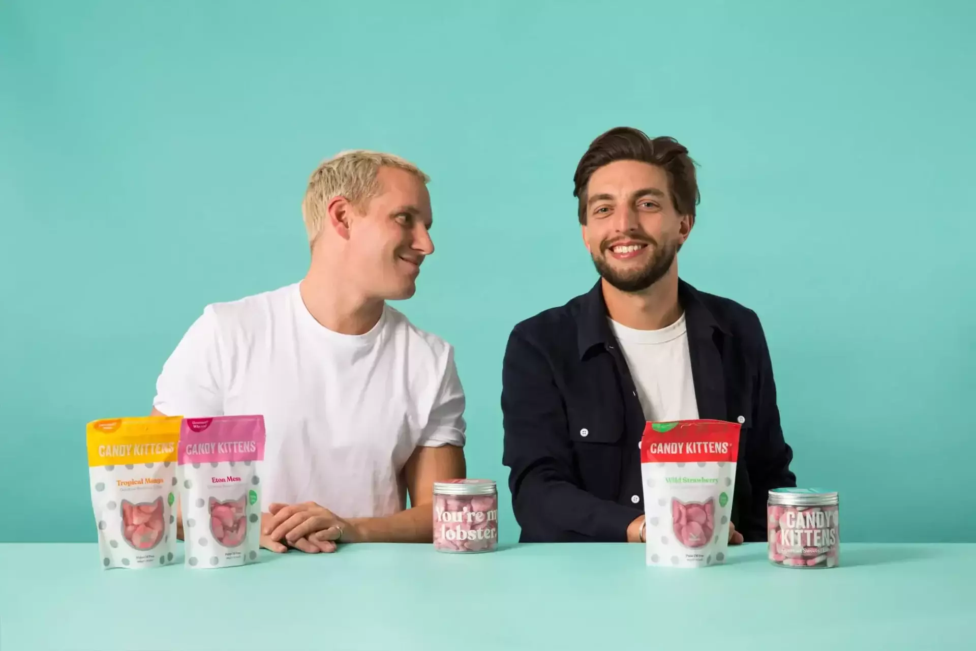 Two men are seated at a table with an array of Candy Kittens products displayed in front of them against a teal background, creating a scene as sweet as the candy itself.