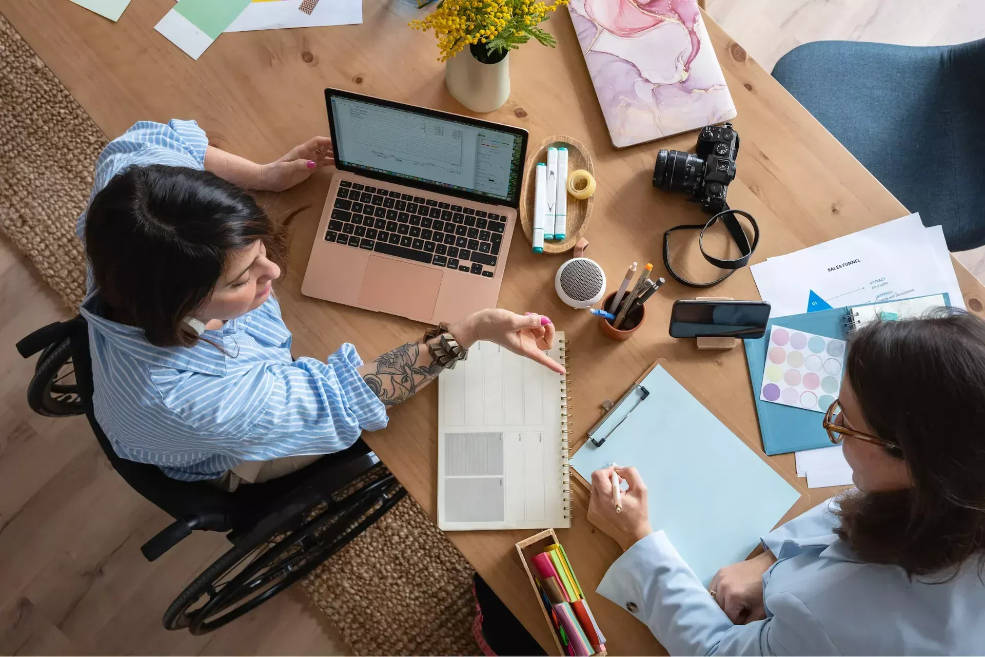 Two women work together at a desk with a laptop, documents, and camera—collaborating on projects for marketing agencies; one woman uses a wheelchair.