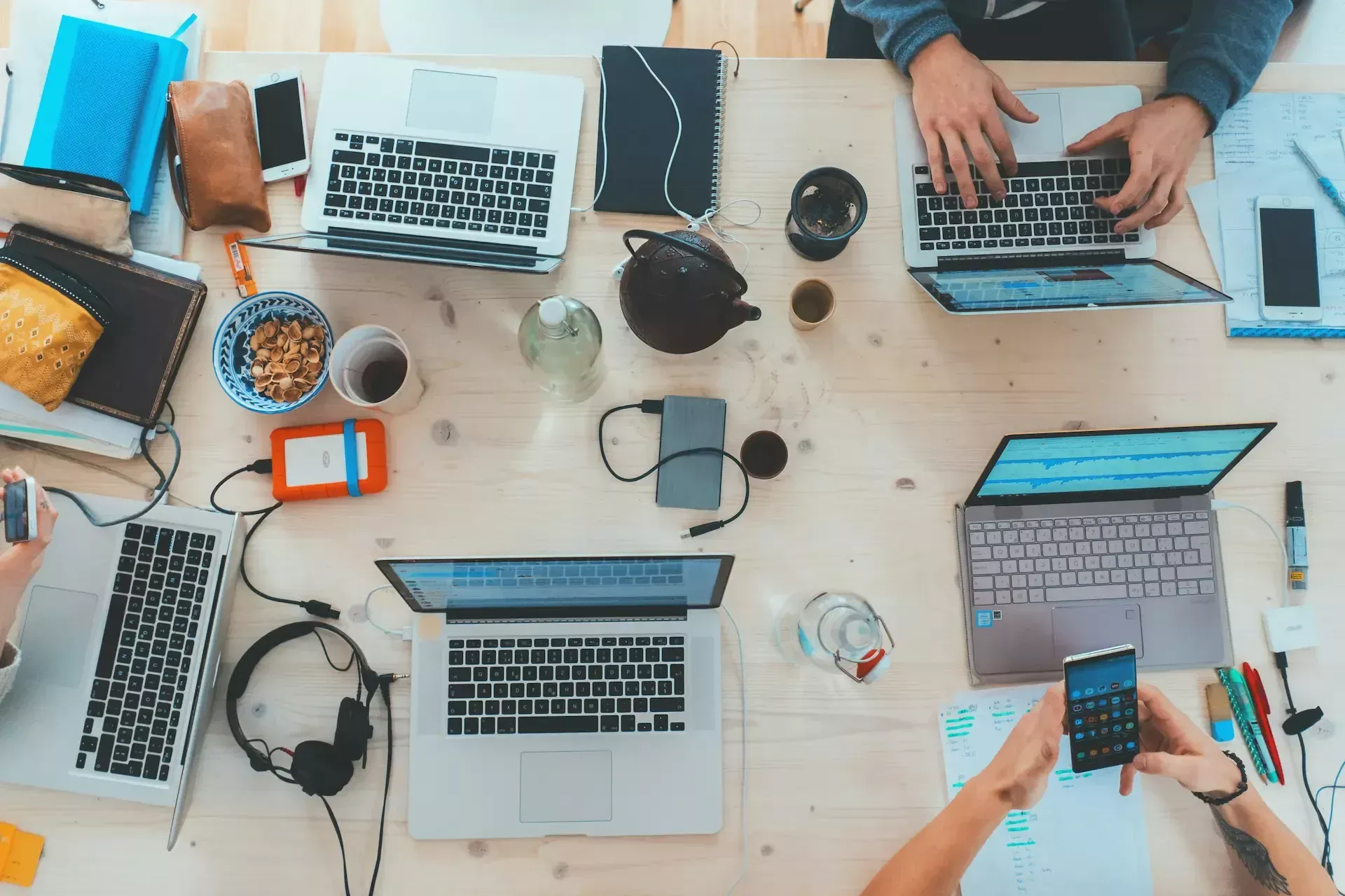 A group of people work at a table with laptops, notebooks, phones, snacks, and office supplies spread across the surface.