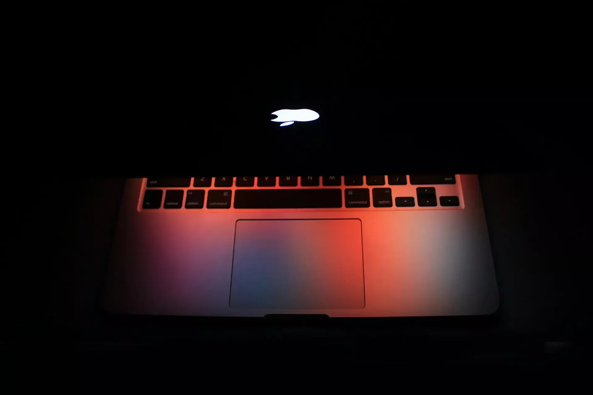 A partially closed macbook laptop with an illuminated apple logo casts red and blue light on the keyboard and trackpad in a dark environment, hinting at concerns around cyber breaches and the growing importance of a cybersecurity budget.