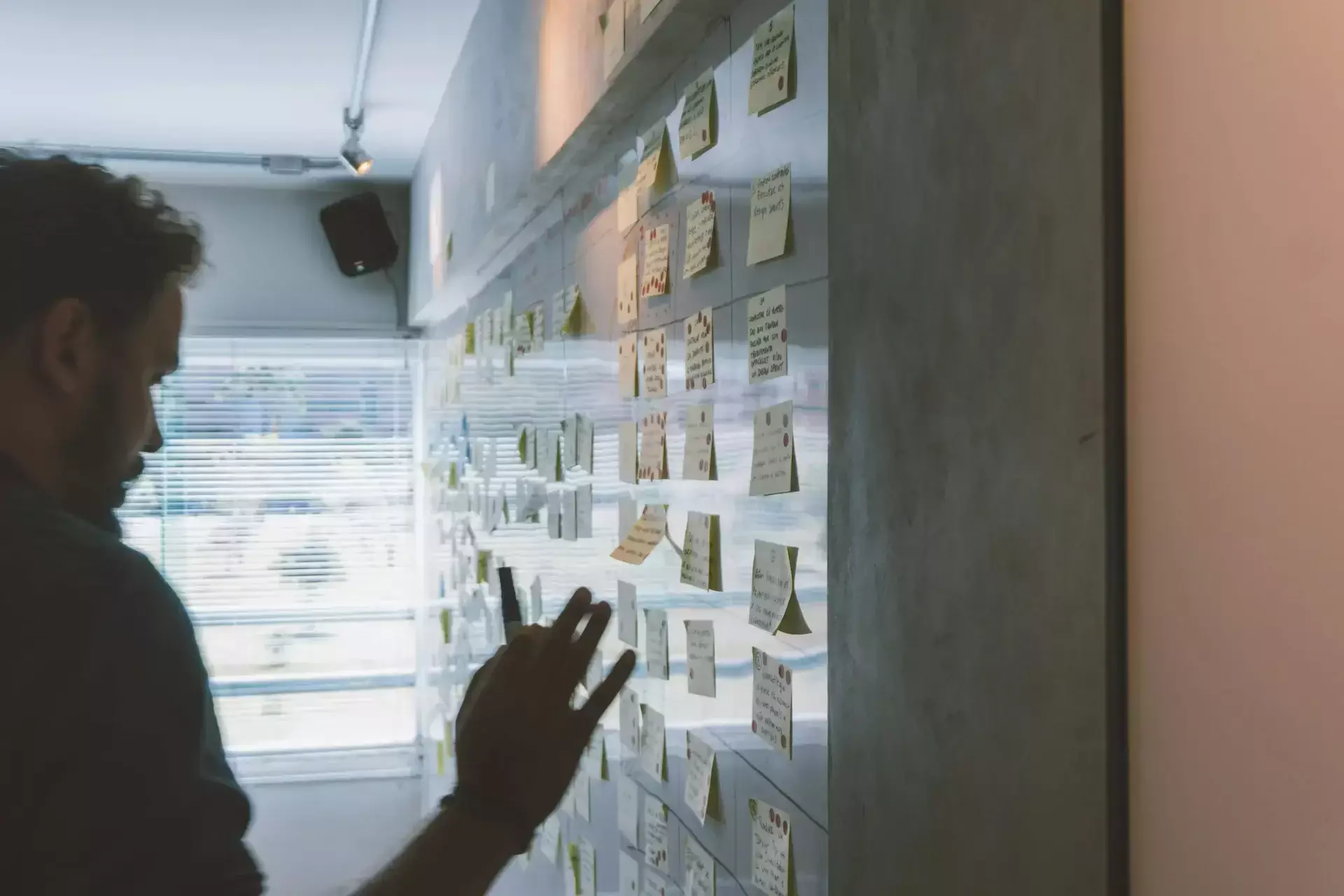 A person stands in front of a wall covered with numerous sticky notes, some written on, in a brightly lit room with large windows, preparing for quarterly business reviews led by the virtual cio.