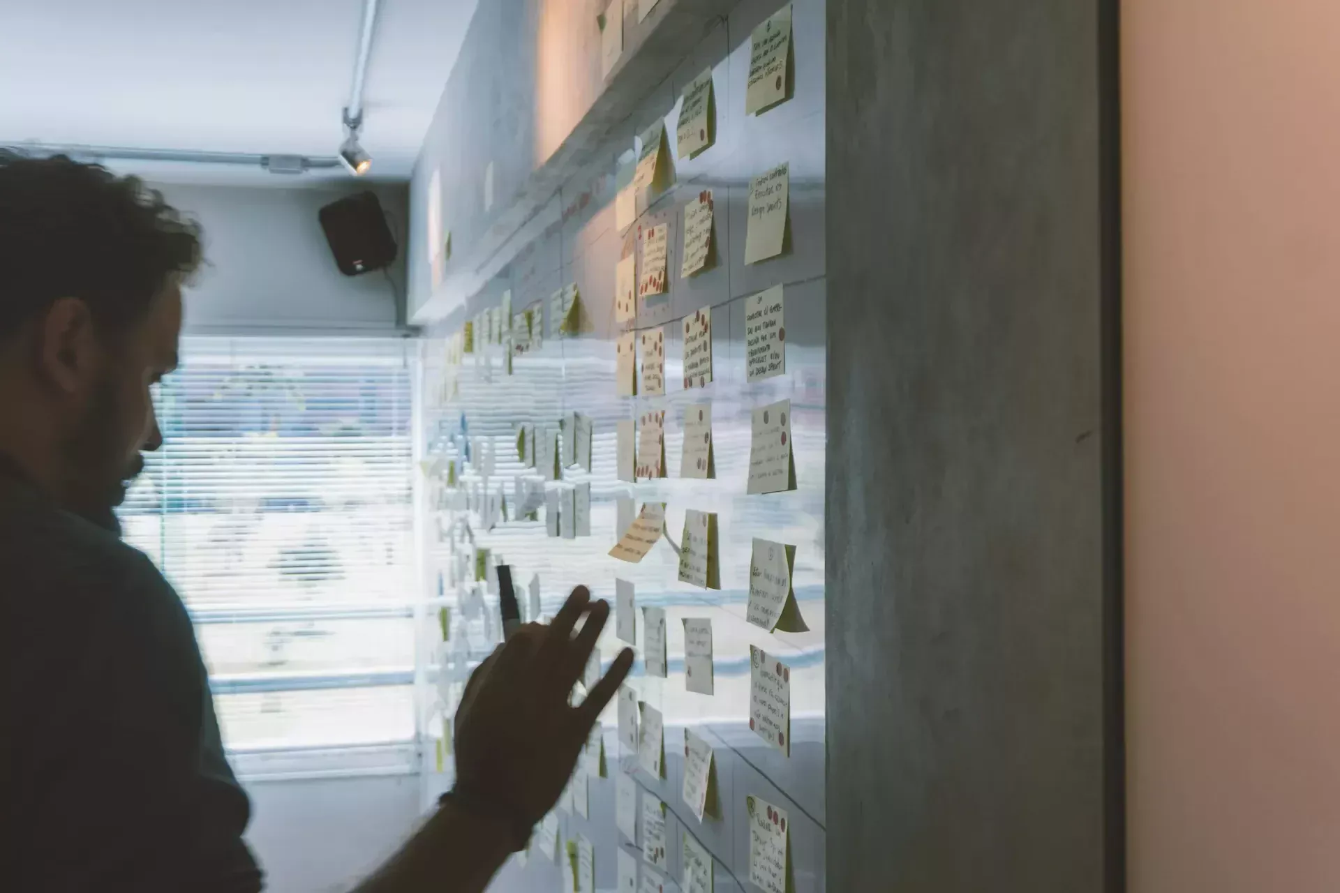 A person stands in front of a wall covered with numerous sticky notes, some written on, in a brightly lit room with large windows, preparing for quarterly business reviews led by the virtual cio.